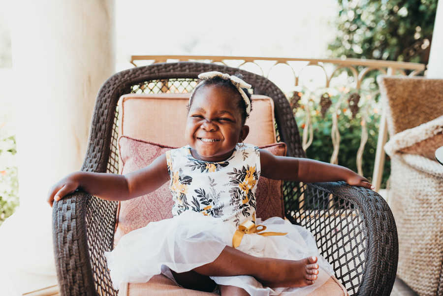 Adopted toddler from Ghana with HIV sits smiling in wicker chair with dress on
