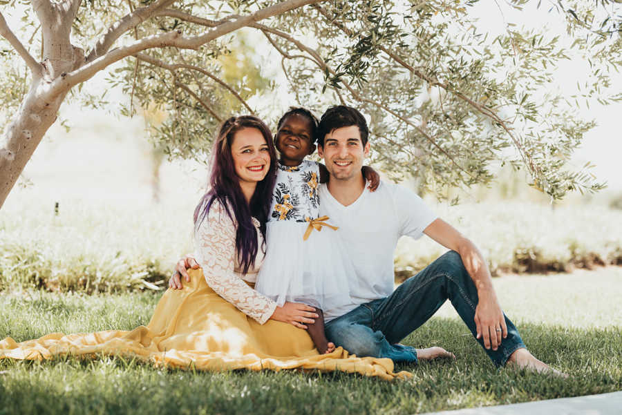 Husband and wife sit on grass smiling with their adopted daughter standing between them