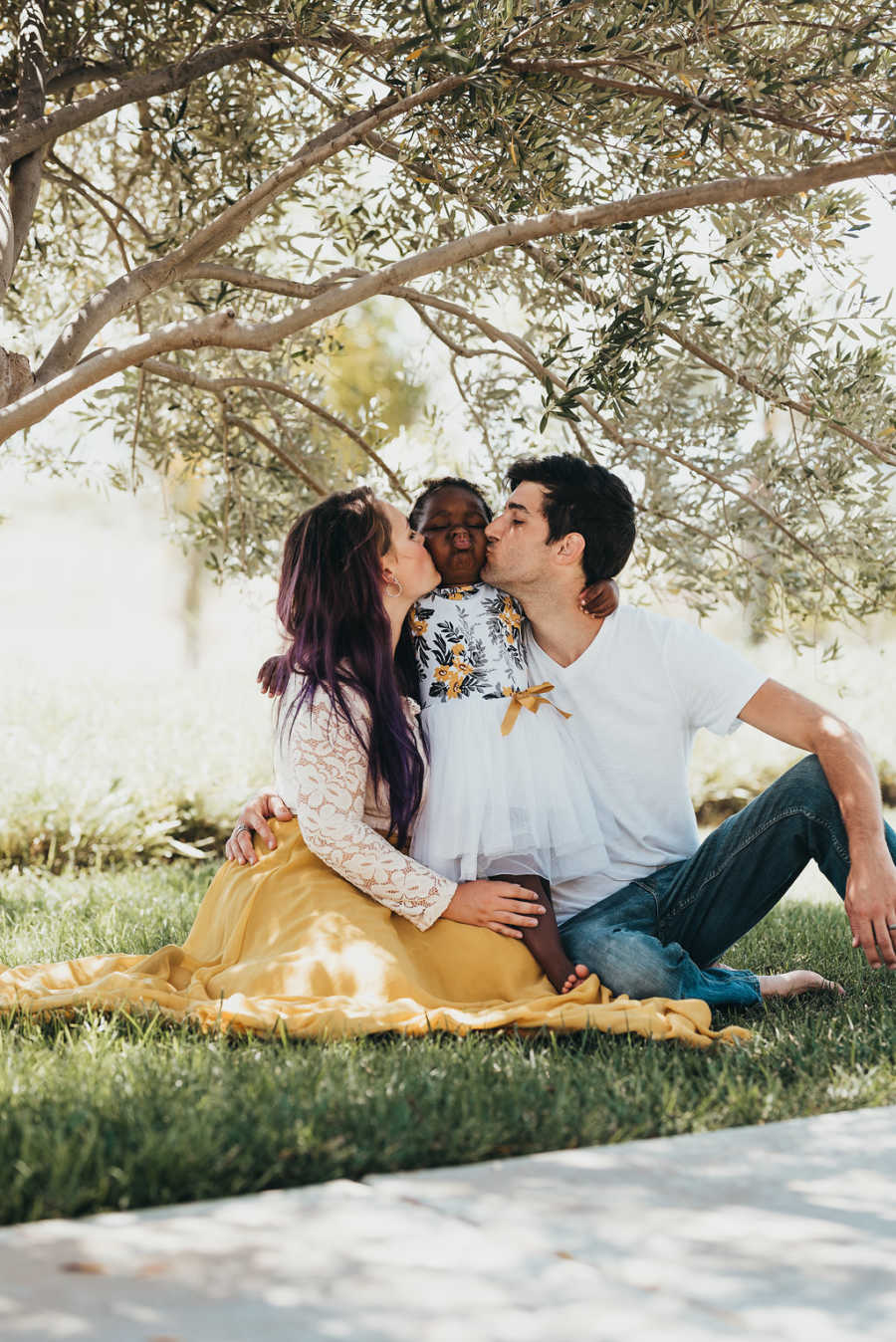 Husband and wife sit on grass kissing their adopted daughters cheeks who sits between them