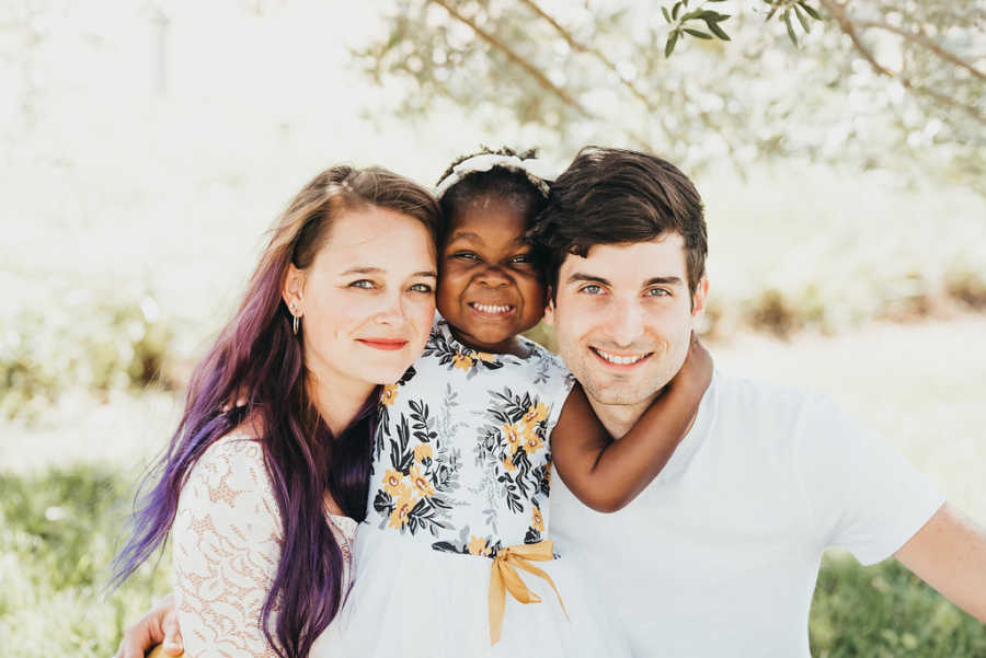 Husband wife and their adopted daughter smile for photoshoot in grassy area