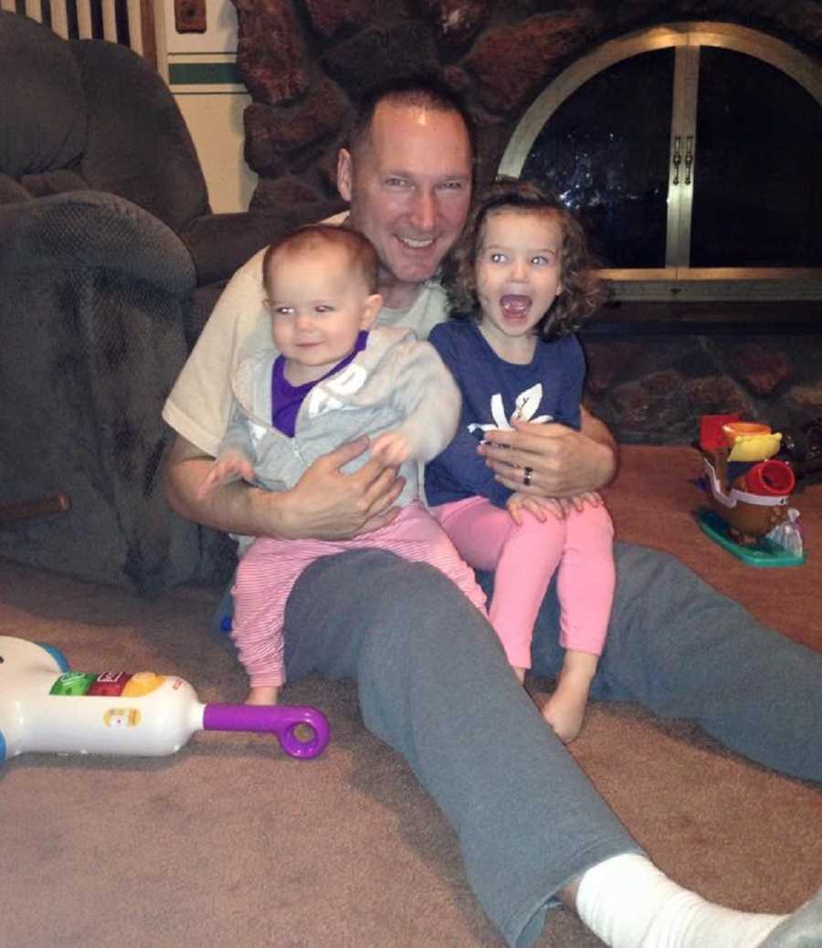 Grandfather sits on floor leaning against chair with two grandchildren sitting in his lap