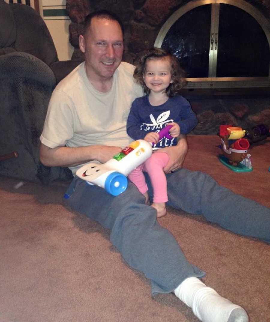 Man sits on floor smiling with granddaughter and her toy in his lap