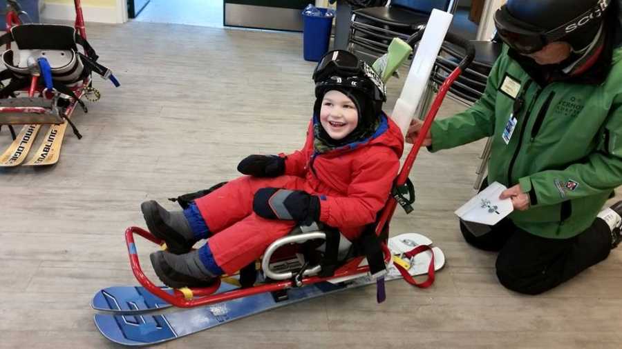 Boy with vanishing white matter disease smiles while strapped into seat on snowboard