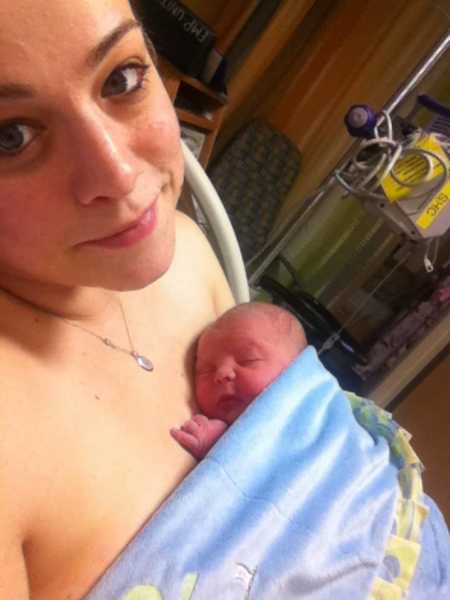 Woman lays in hospital bed smiling in selfie with newborn son sleeping on her bare chest