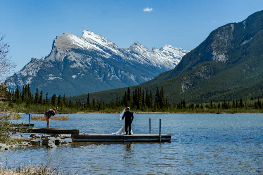 Bride whose chances to live were against her stands on dock with husband next to body of water and mountains