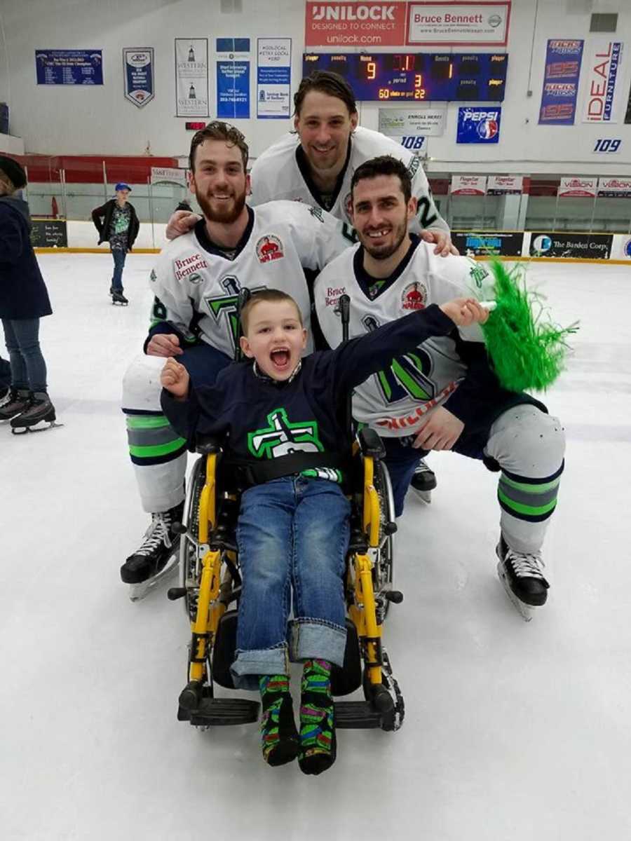 Boy with vanishing white matter disease sits in wheelchair on ice rink with three hockey players smiling behind him