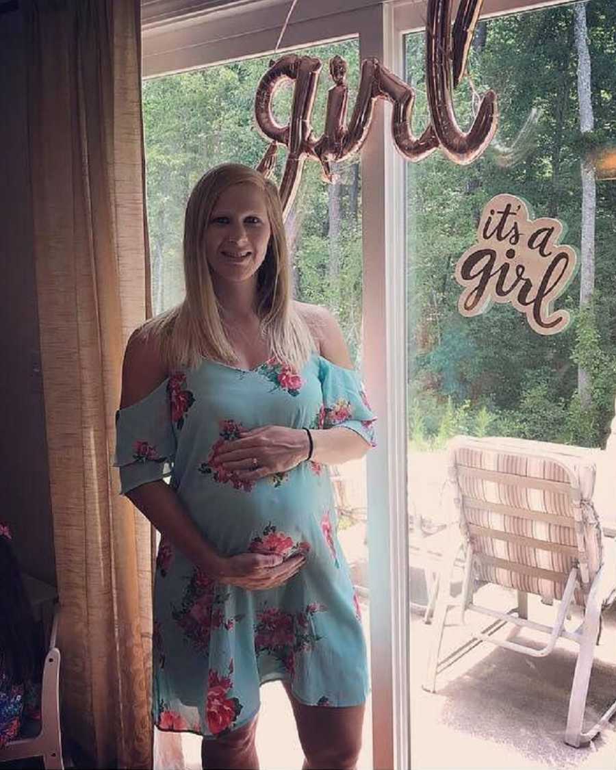 Pregnant woman stands holding her stomach by glass door with ballon saying "girl" and sign saying, "It's a girl"