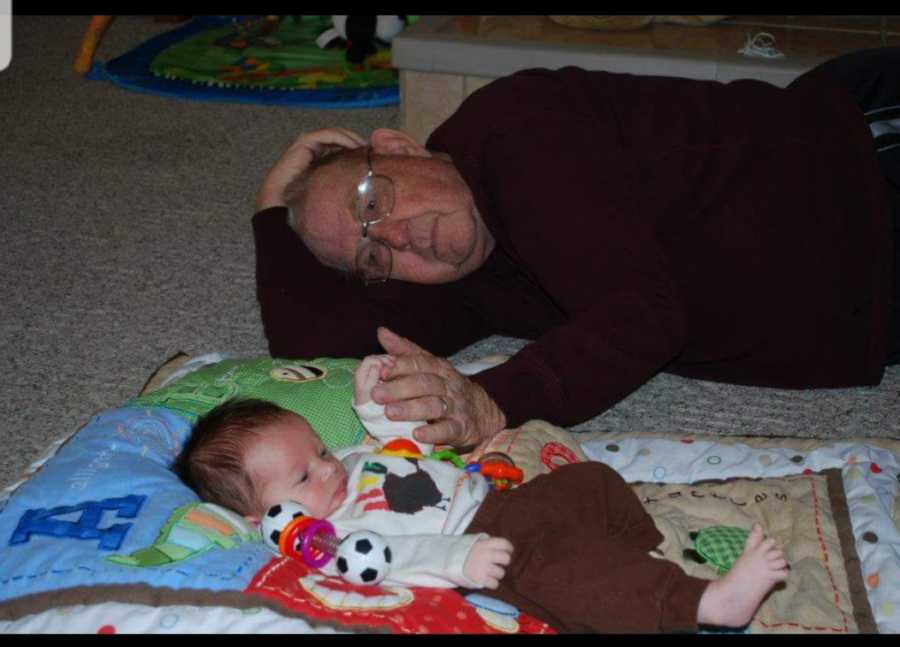Baby boy lays on blankets on the ground with his grandpa laying beside him holding his hand