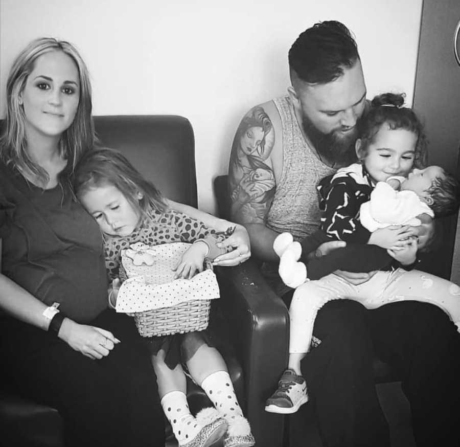 Mother sits in chair with her daughter beside husband who has their other daughter in his lap who holds newborn