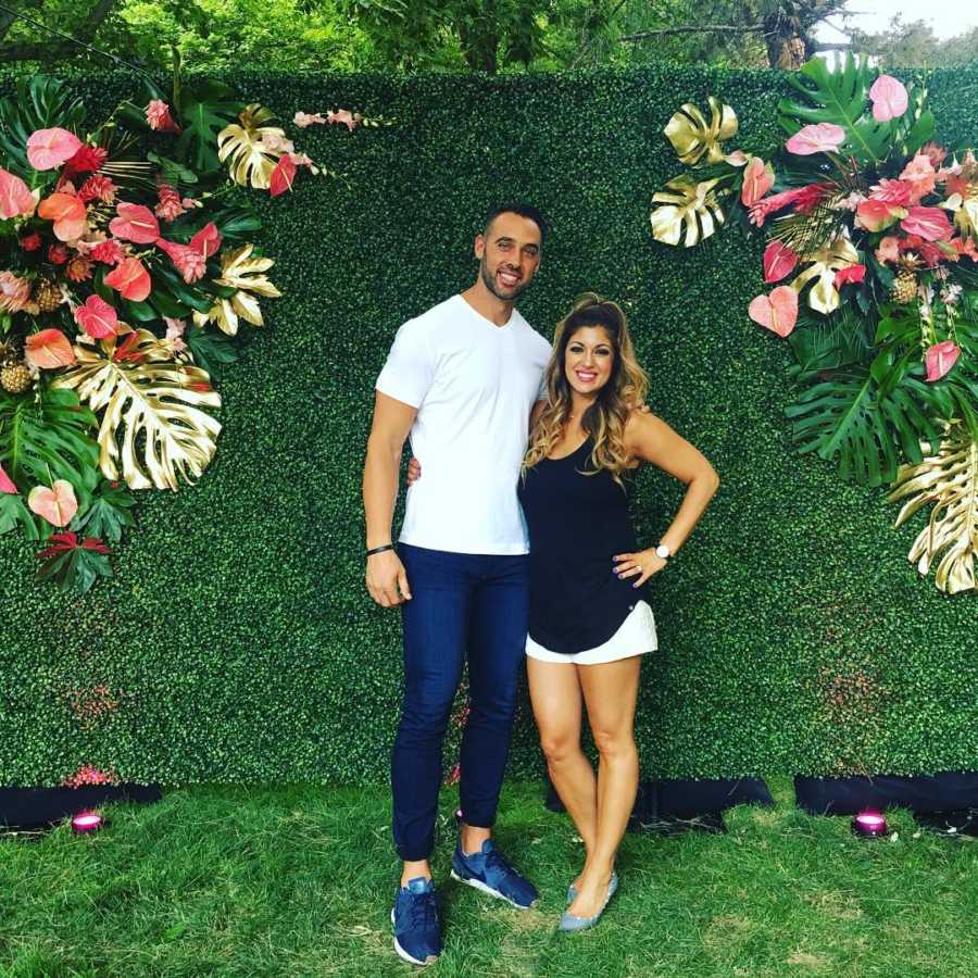 Married couple stand smiling in front of turf backdrop with tropical flowers 