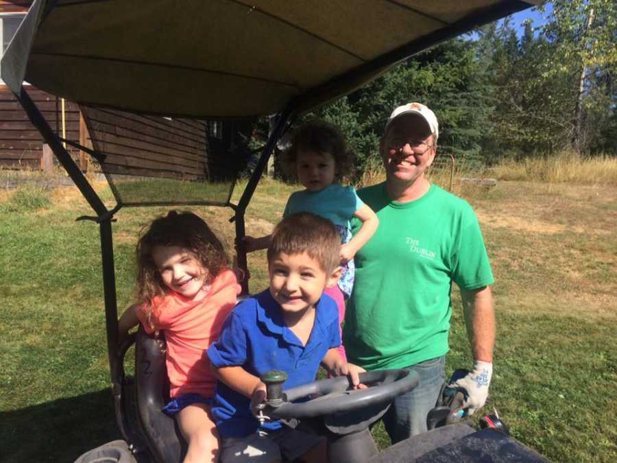 Grandfather stands beside three grandchildren who are sitting in his sit down lawn mower