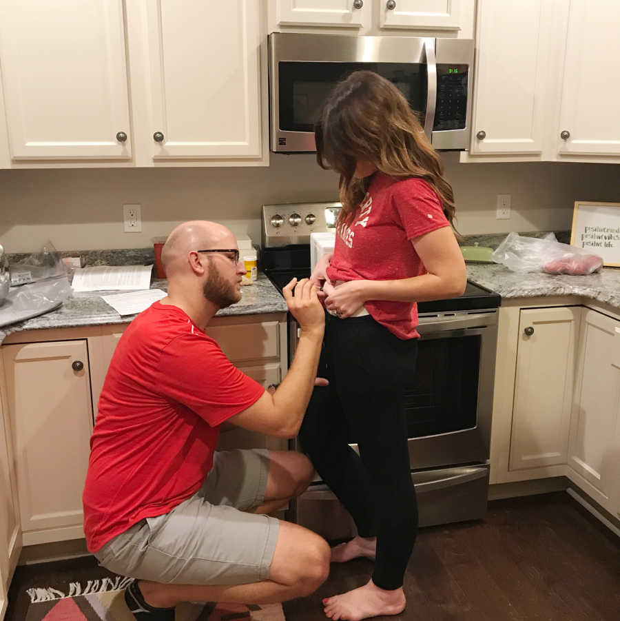 Husband kneeling in front of wife's stomach injecting IVF needle in their kitchen