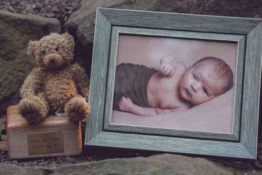 Teddy bear sitting on box of ashes of infant who died from SIDS besides picture frame of him
