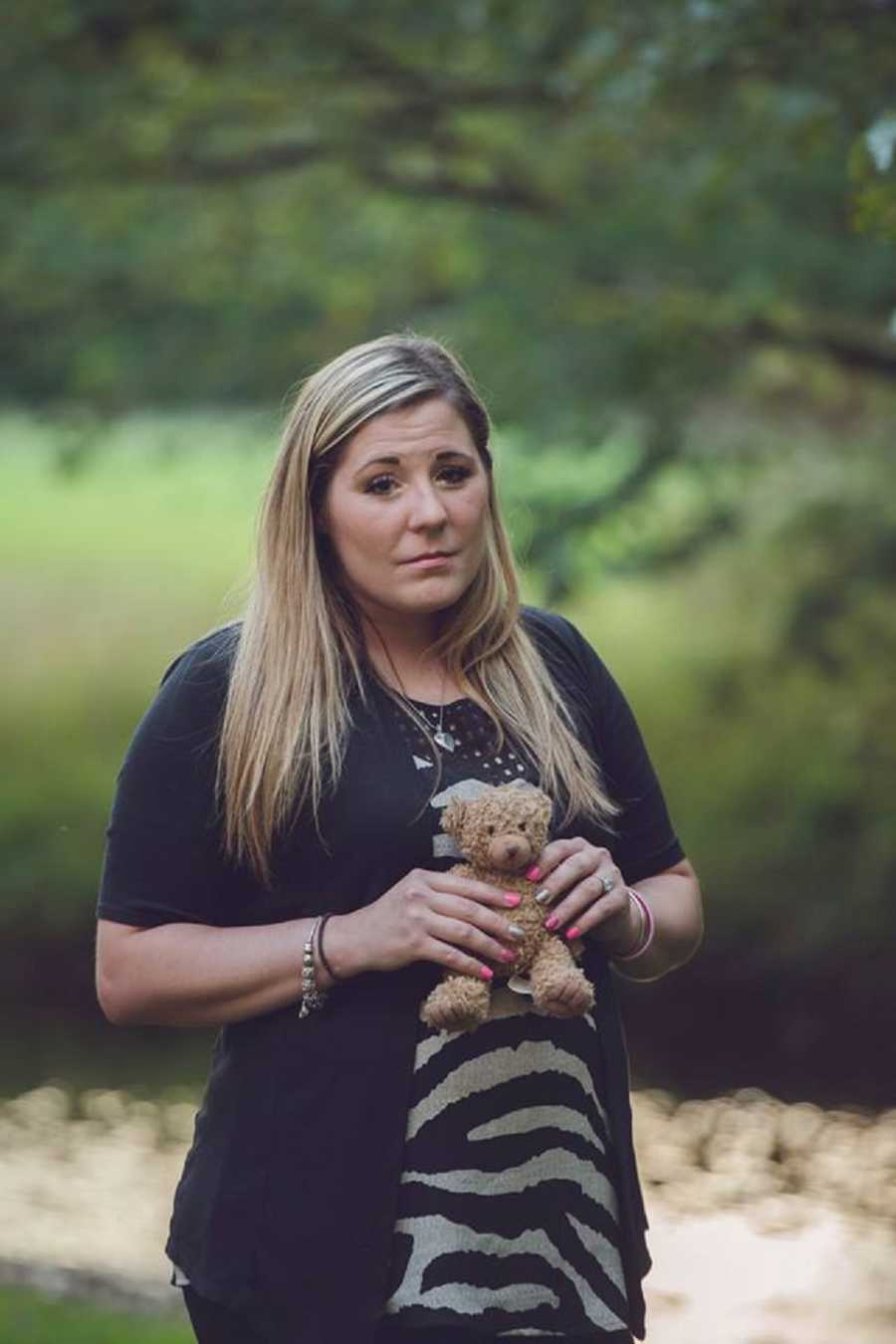 Mother who lost her infant to SIDS stands frowning with little teddy bear