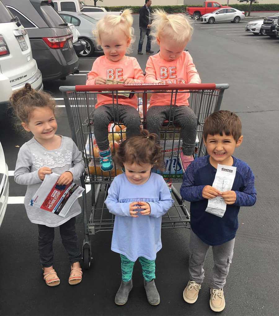 Twins sit in grocery cart in parking lot with three older siblings standing in front of cart