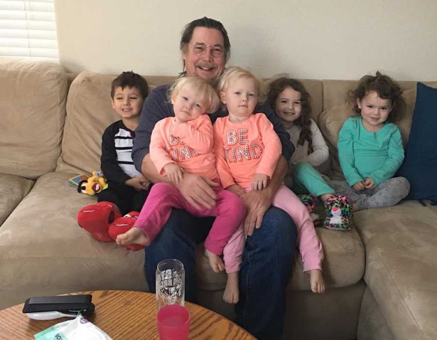 Grandfather sitting on couch with twins in his lap and three other grandchildren on either side of him