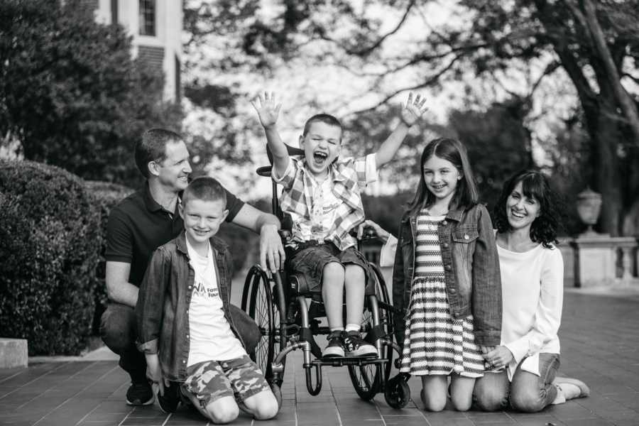 Boy with vanishing white matter disease sits in wheel chair with hands in the air beside parents, brother and sister