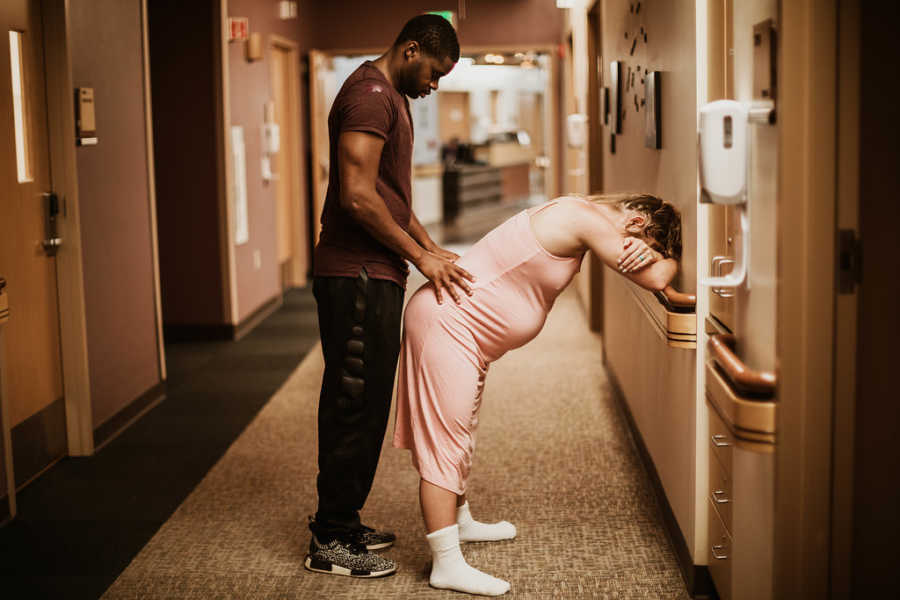 Pregnant woman leans over leaning against hospital wall while husband massages her back