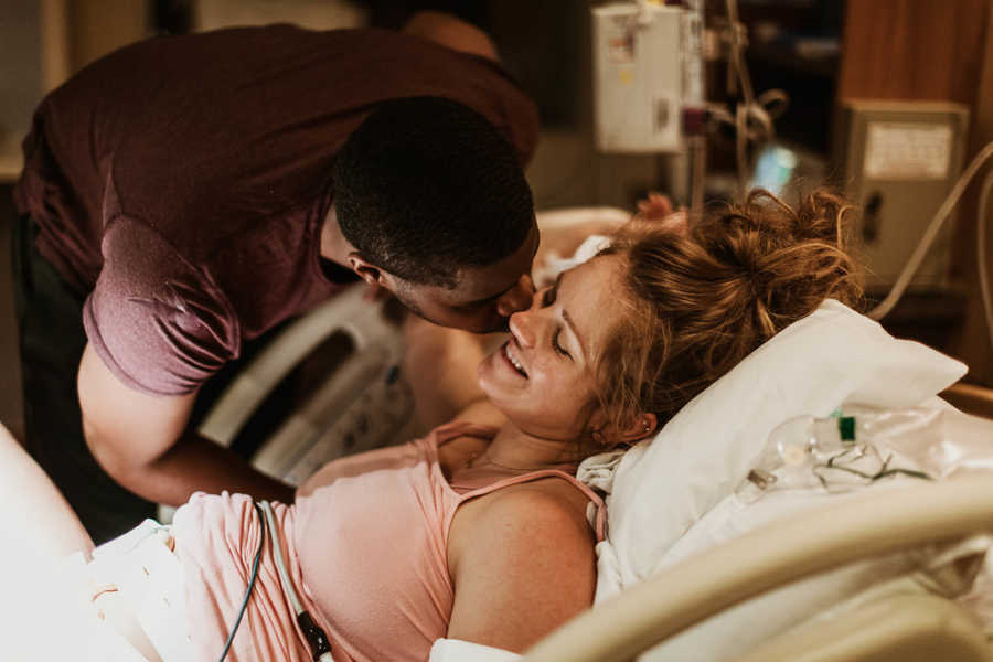 Man leans over hospital bed to kiss his wife on the cheek who just gave birth