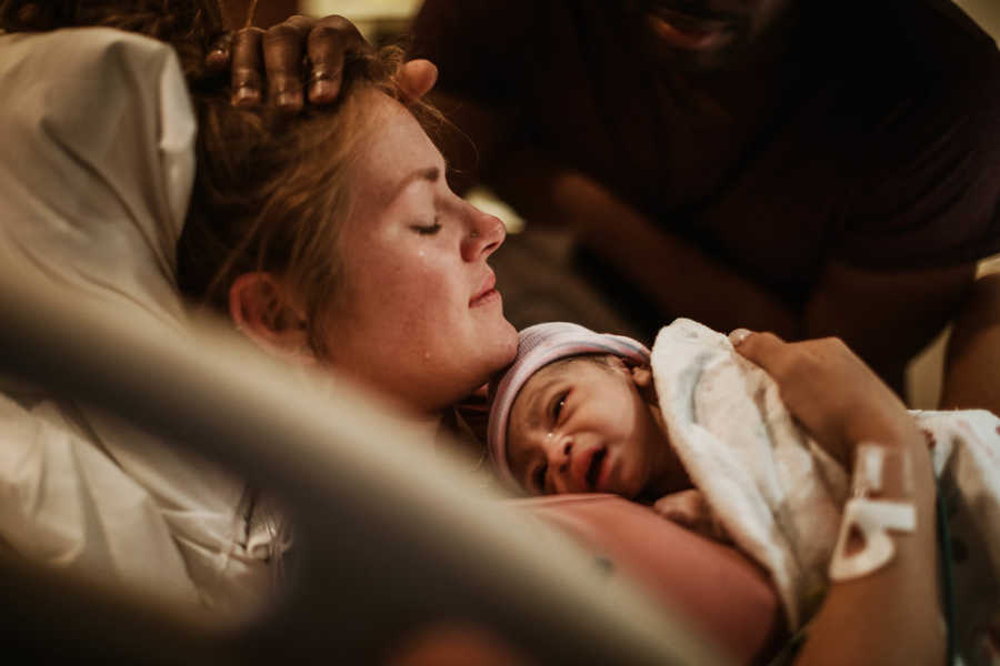 Mother holds newborn in hospital bed to her chest while husband rests hand on wife's head