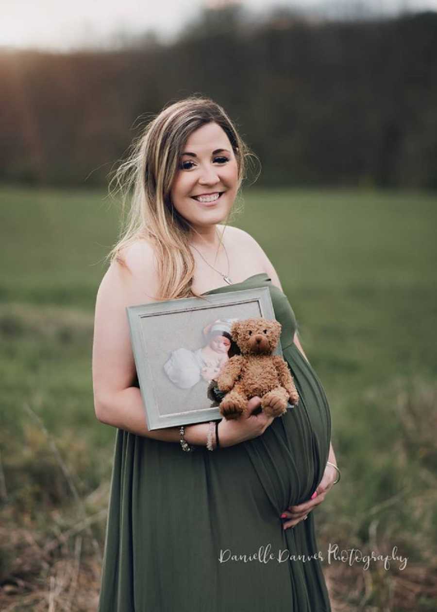 Pregnant woman who lost her first born to SIDS stands smiling holding her stomach with picture of first born