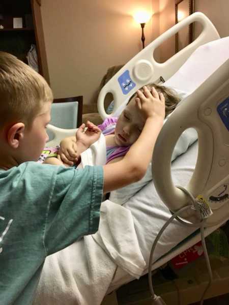 Older brother stands beside sister in hospital bed holding his hand to her forehead