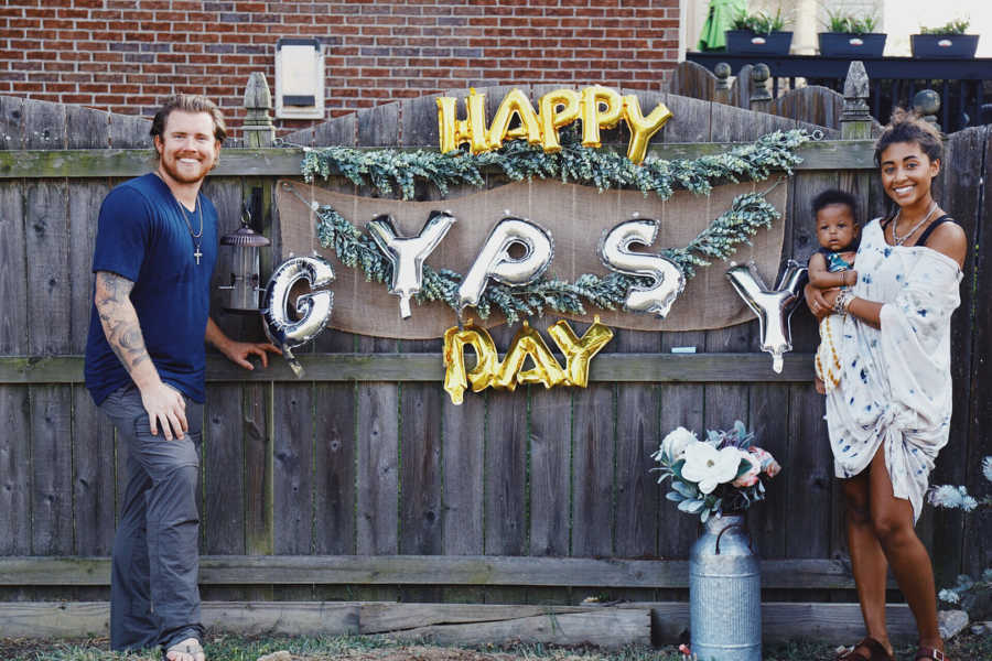 Mother holds adopted daughter on one end of balloon sign that says, "Happy Gypsy Day" with husband on other end