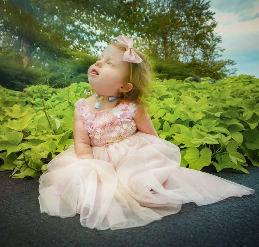 Little girl with Trisomy 18 sitting on sidewalk in pink dress looking up at the sky