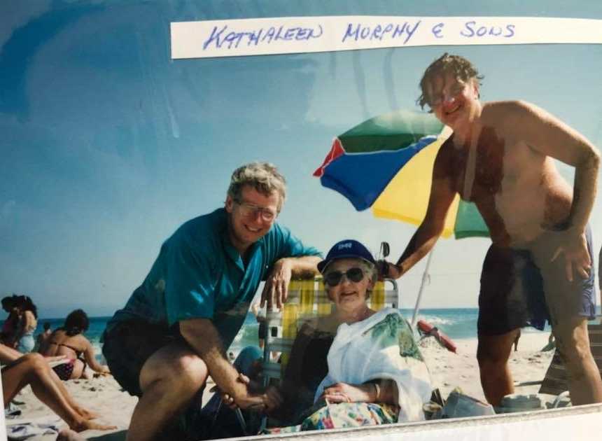 Elderly woman sitting in beach chair with one man squatting beside her and another standing over her