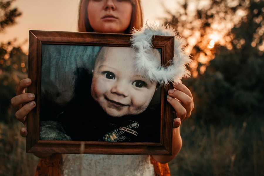 Mother holds out picture frame of son who passed from SIDS with feather halo resting on corner of frame