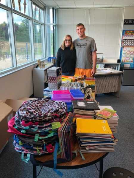 Teen stands with arm around teacher behind table filled of school supplies he donates to kids