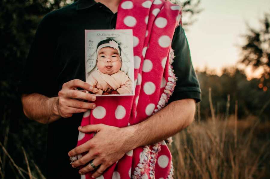 Father holds picture of deceased newborn with her pink polka dot blanket on his shoulder