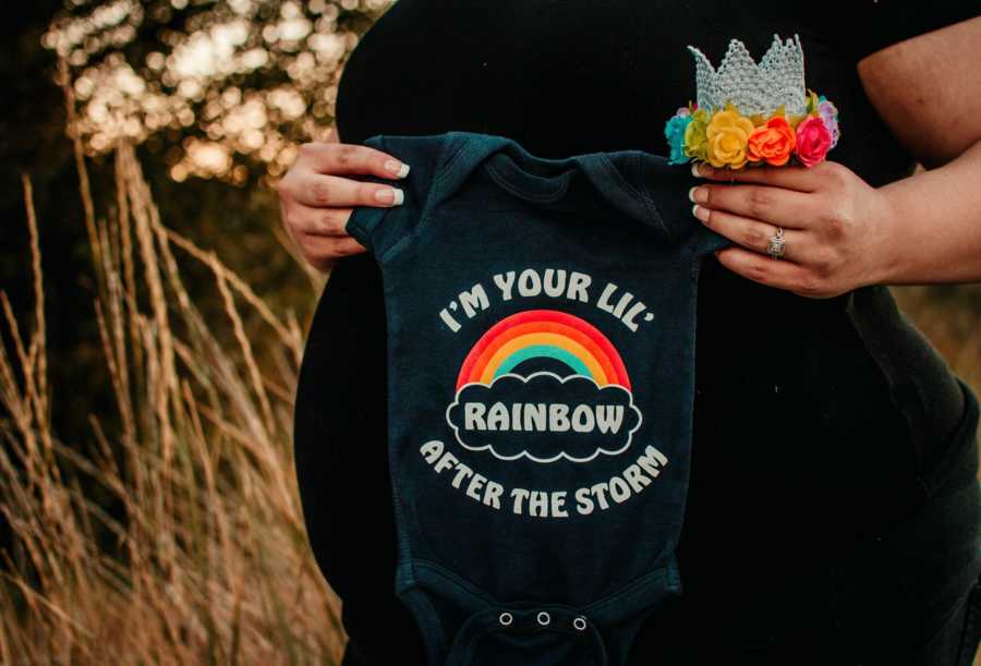 Pregnant mother who lose her first child holds onesie that says, "I'm your lil rainbow after the storm" 