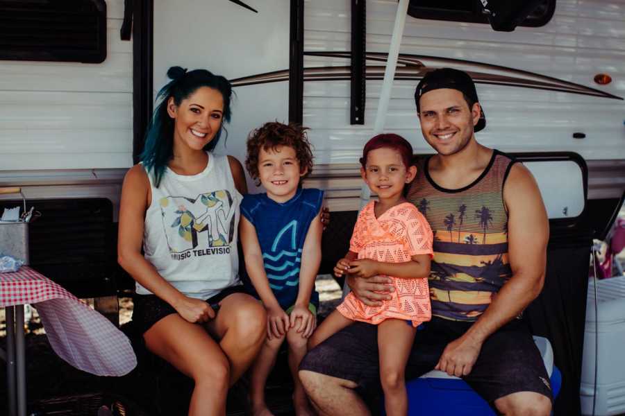 Husband and wide sit smiling in front of their RV with their son and daughter in between them