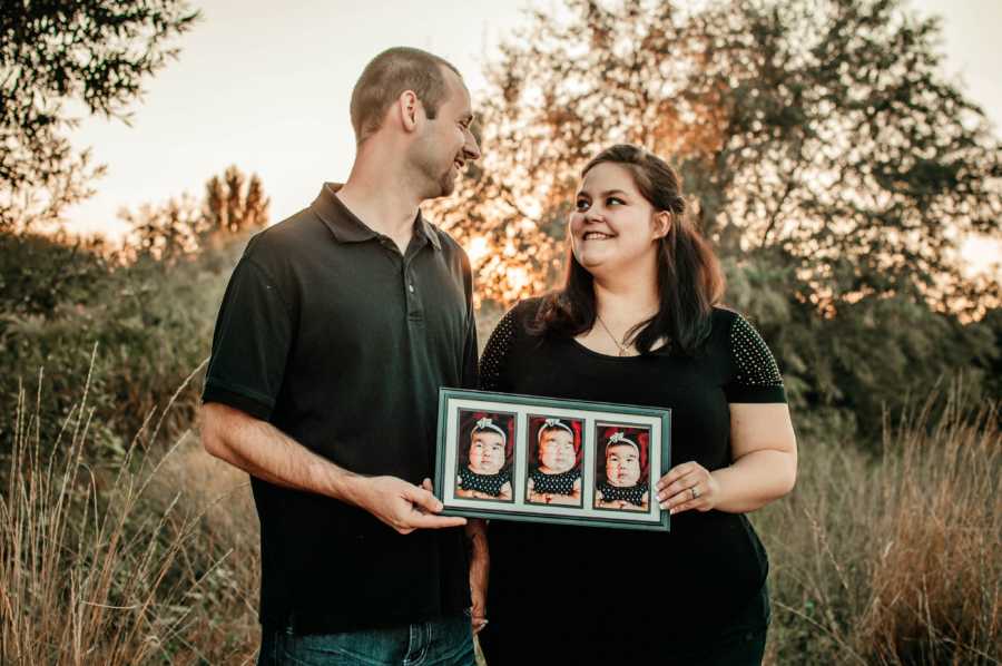 Husband and wife stand in field holding picture frame of their deceased newborn