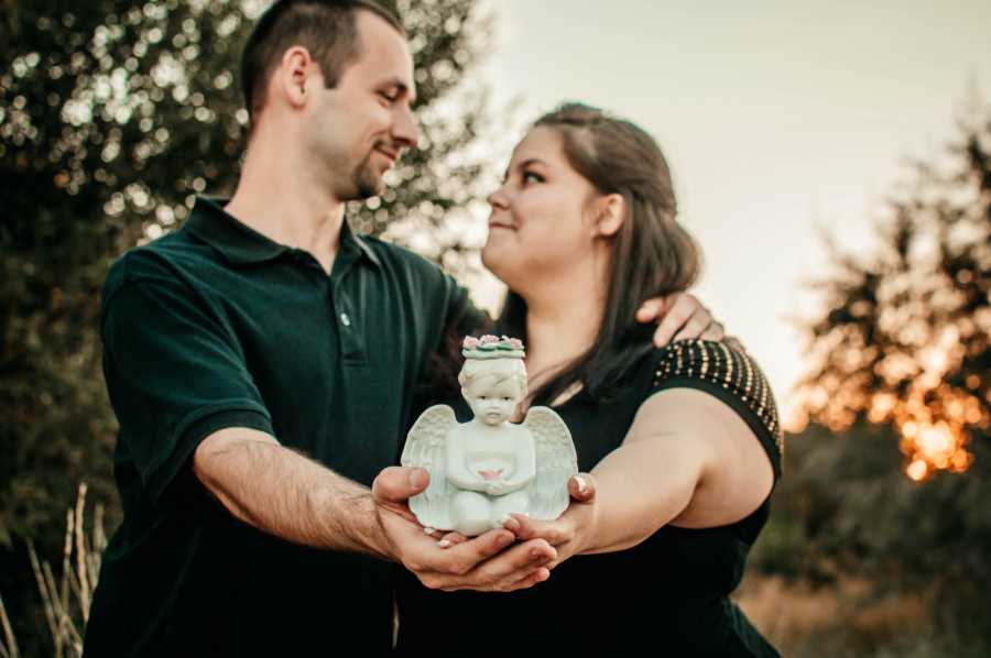 Husband and wife stand arm in arm looking at each other while they hold small angel statue for their decease first born