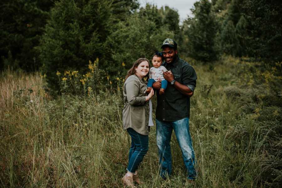Wife stands in field holding infant daughter with husband who is a teacher