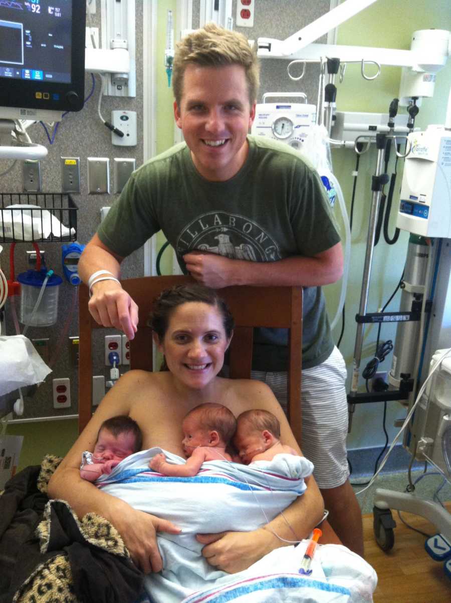 Husband stands behind wife who sits in chair holding triplets to her chest