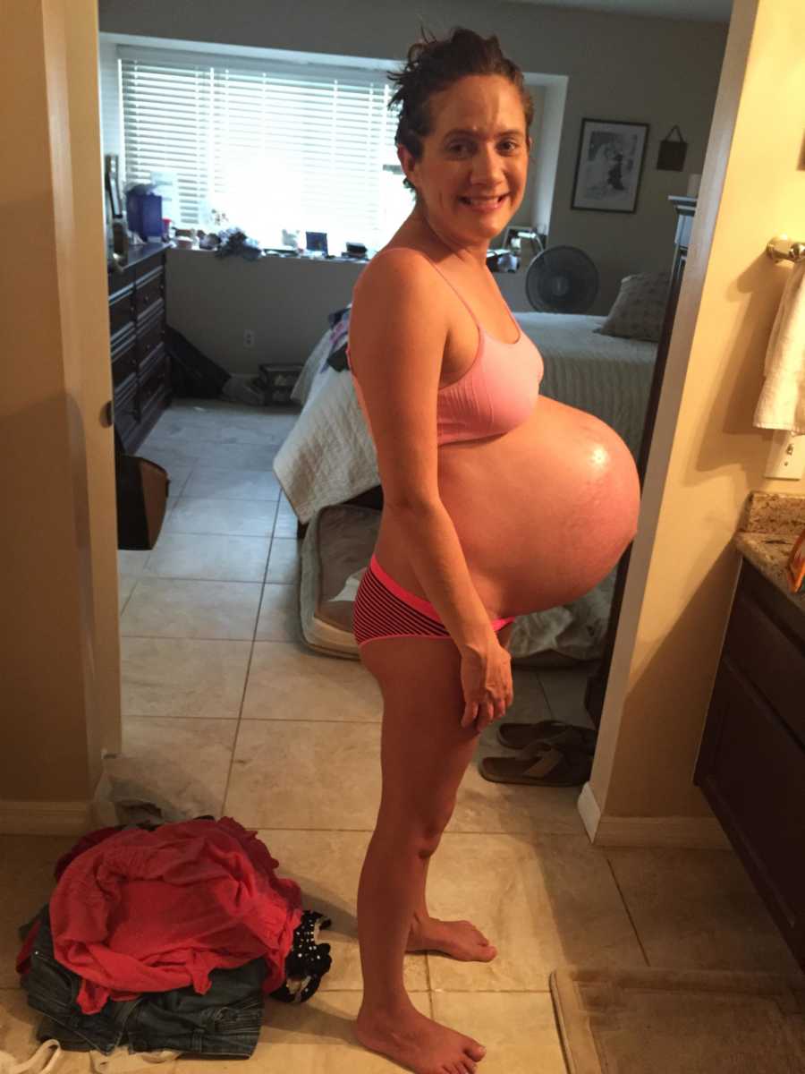 Pregnant woman stands smiling in bathroom wearing bra and underwear