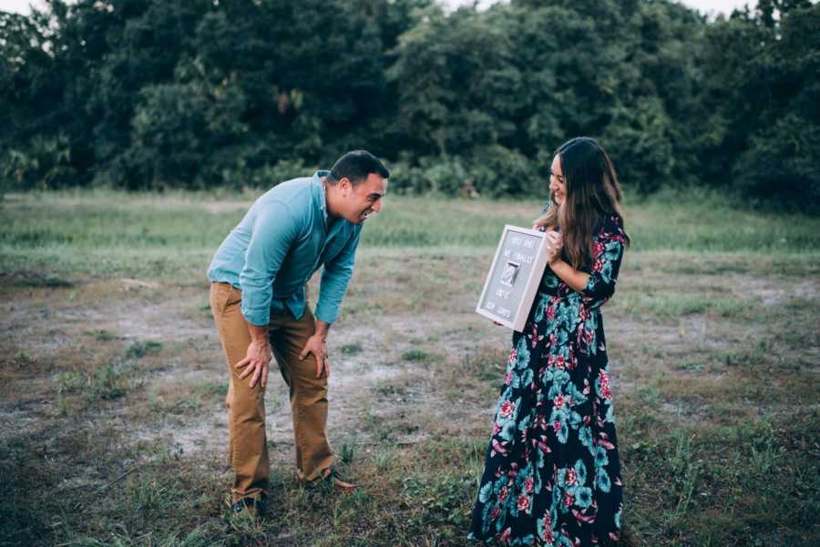 Man rests his hands on his knees as he looks at sign wife holds announcing her pregnancy