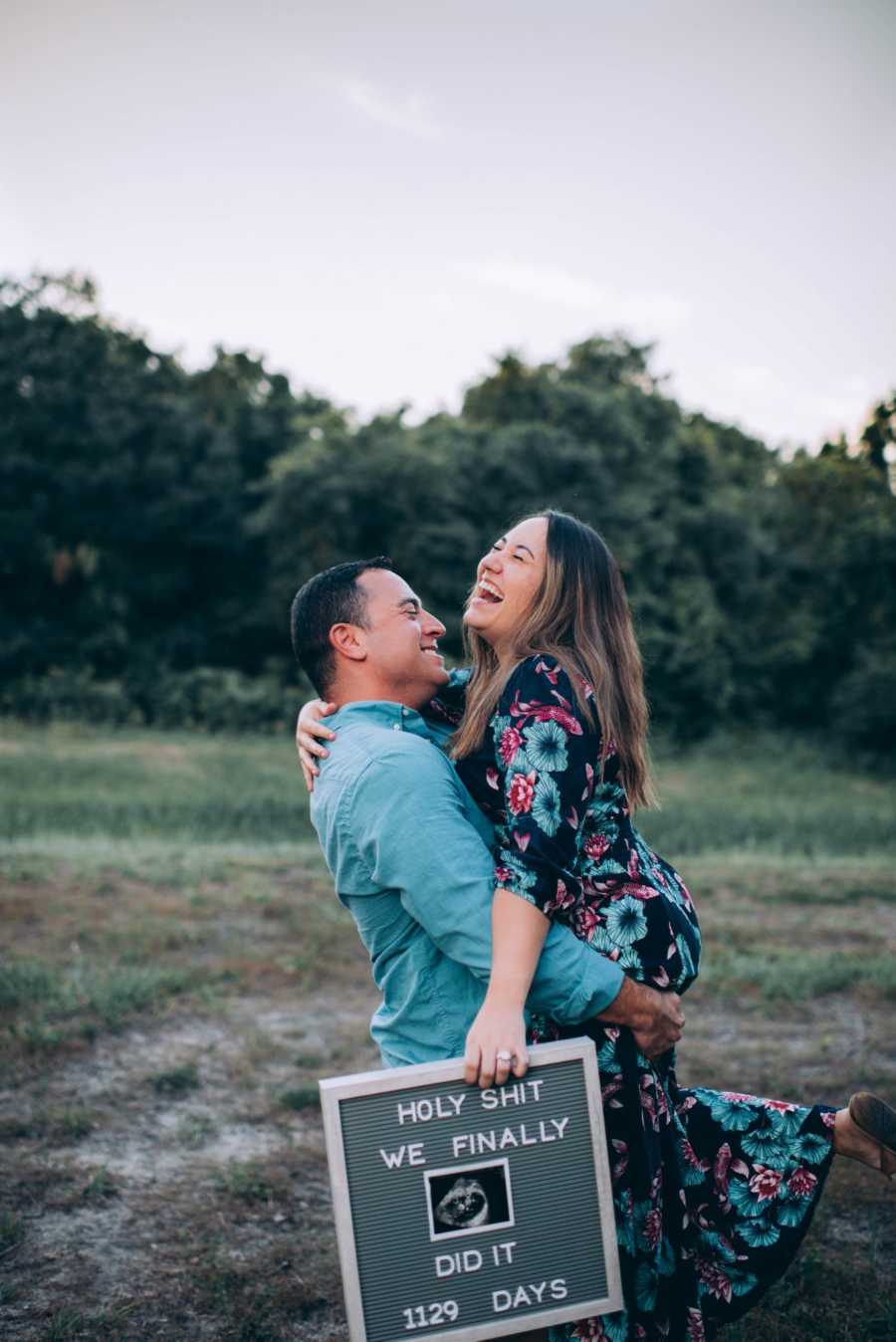 Husband holds up wife who is holding sign announcing her pregnancy after struggling with fertility