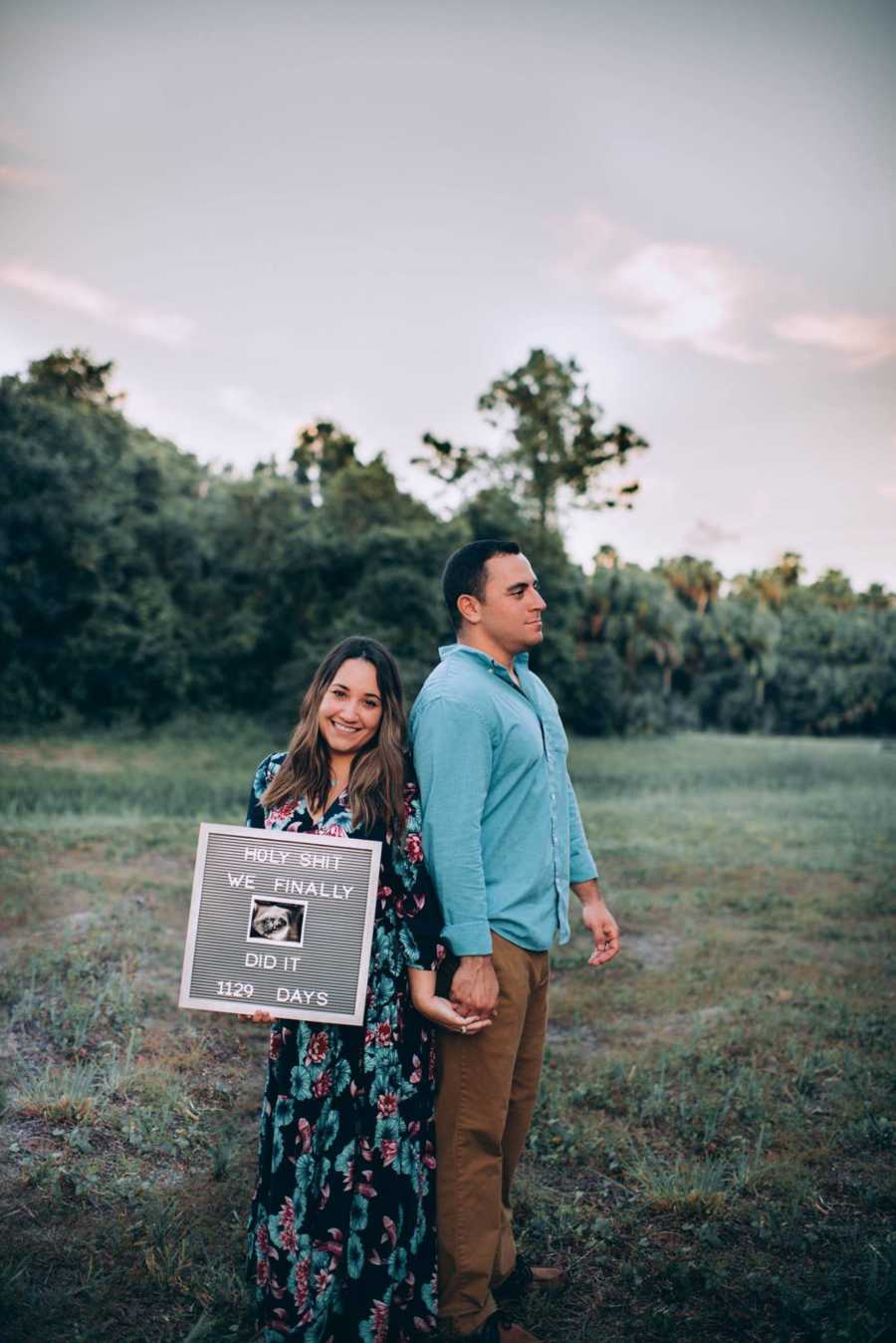 Husband and wife stand back to back holding hands in field while bride holds sign announcing pregnancy