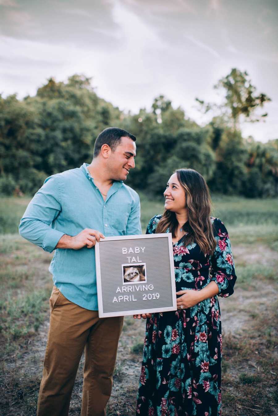Husband and wife stand smiling at each other while holding sign that says, "Baby Tal"