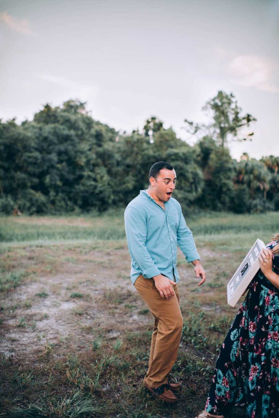 Husband stands in field with jaw dropped looking at sign announcing wife's pregnancy