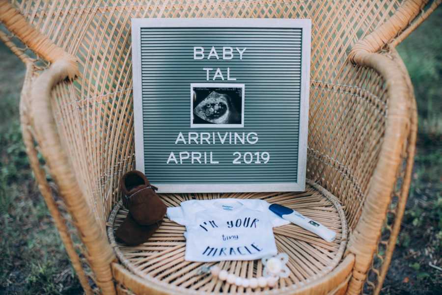 Wicker chair with baby onesie and shoes, pacifier, pregnancy test, and sign with baby's arrival date