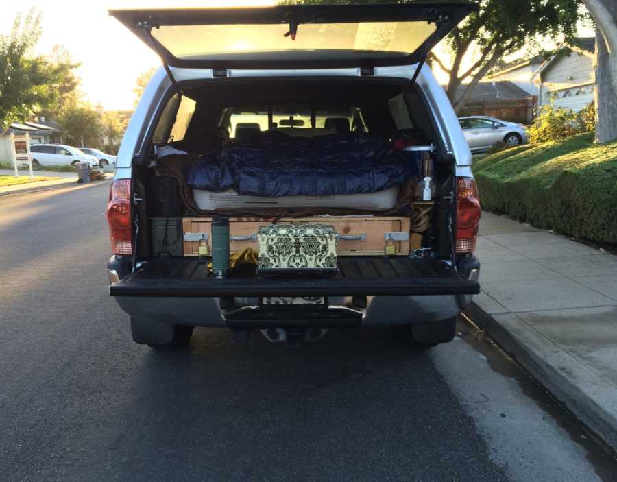 Trunk of car open showing box of wife's ashes in front of blow up bed and comforter