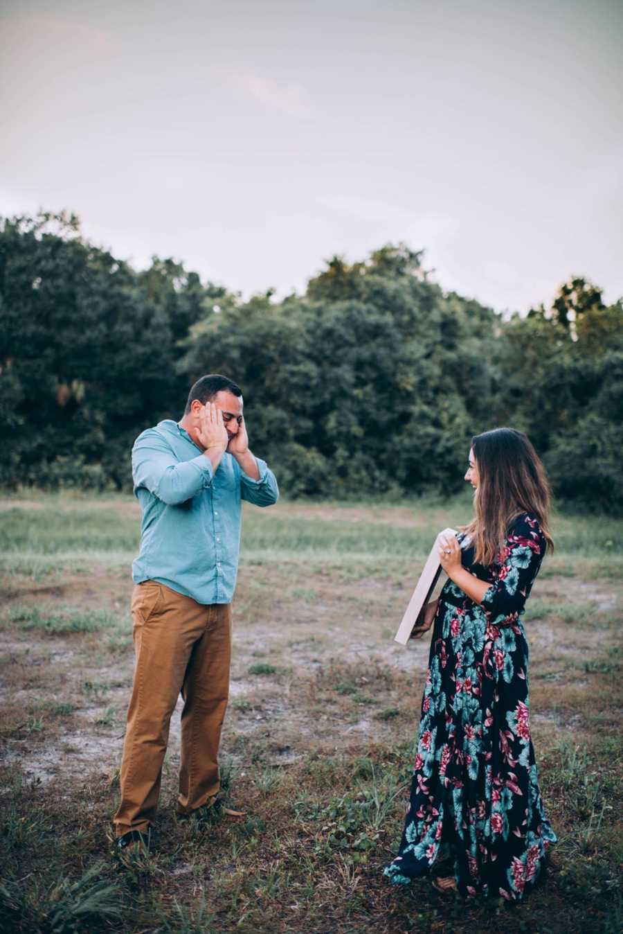 Wife holds up sign announcing pregnancy in front of husband who stands with hands to his head