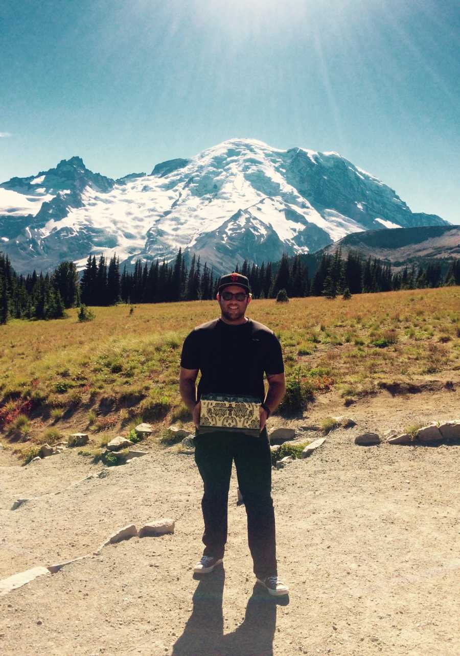 Man stands smiling with box of wife's ashes in his hands with snowy mountains behind him