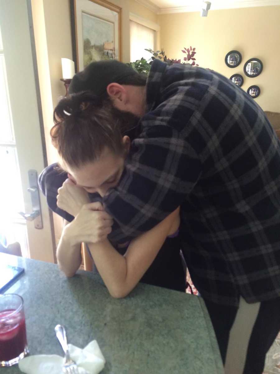 Man stands hugging fiancee who is sitting on stool after hearing she was diagnosed with cancer