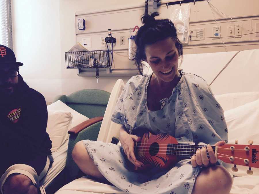 Wife with cancer smiles in hospital bed while playing ukelele with husband beside her